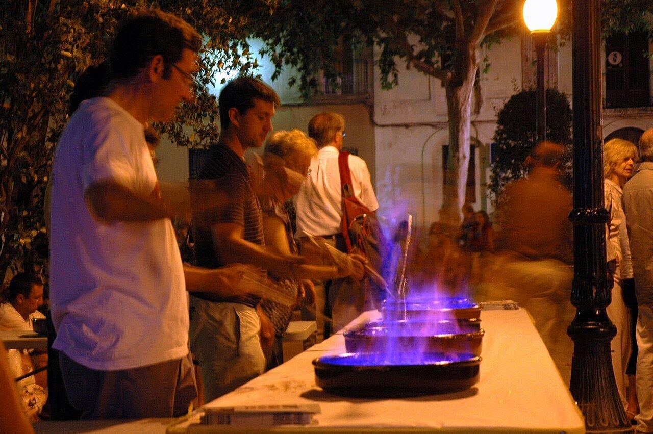 People preparing flaming rum cremat in pans at an outdoor gathering